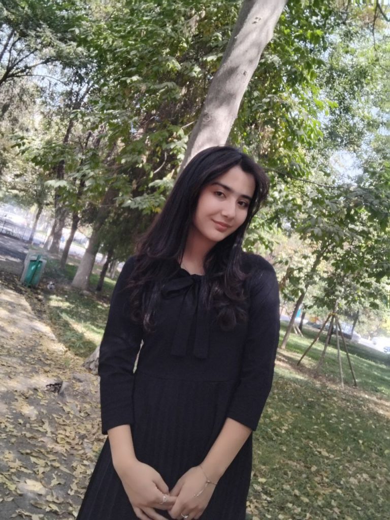 Young Central Asian woman with dark hair and a black outfit outside near green bushes on a sunny day.