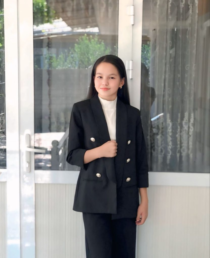 Young Central Asian woman in a black coat, white dress top, and black pants, in front of a window with some green bushes outside. 