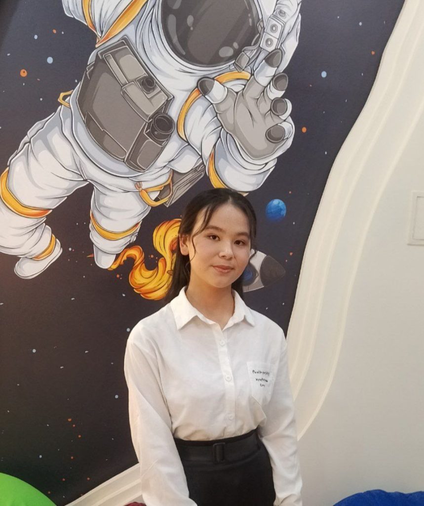 Young Central Asian woman with straight dark hair behind her head and a white collared shirt, standing in front of a photo of an astronaut. 