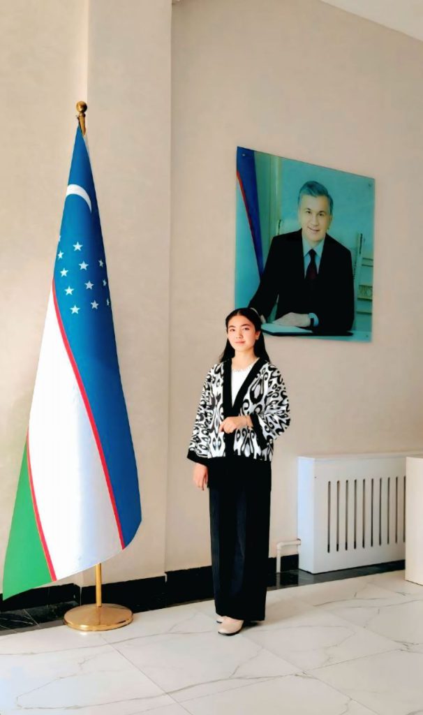 Young Central Asian woman with a black and white patterned coat, black pants, and long dark straight hair. She's standing near the green, white, and blue Uzbek flag and near a photo of a person in a suit. 
