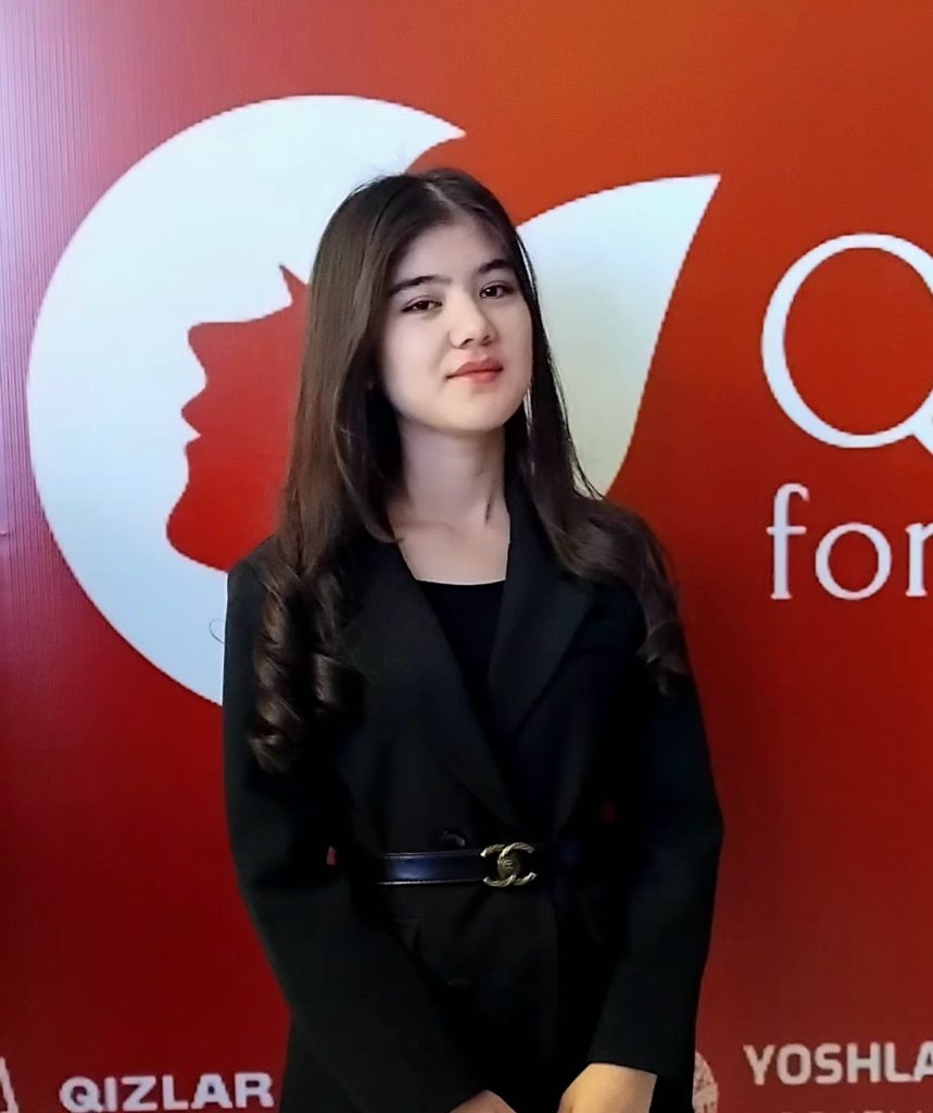Young Central Asian woman with long curly dark hair, brown eyes, a black coat, standing in front of a red sign background. 