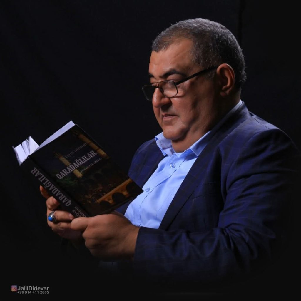 Older middle ages Central Asian man in a dark blue coat over a light blue collared shirt reading a book.