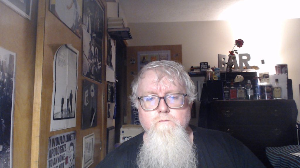 Middle aged white man with reading glasses and a long beard standing in a bedroom with posters on the wall and a dresser