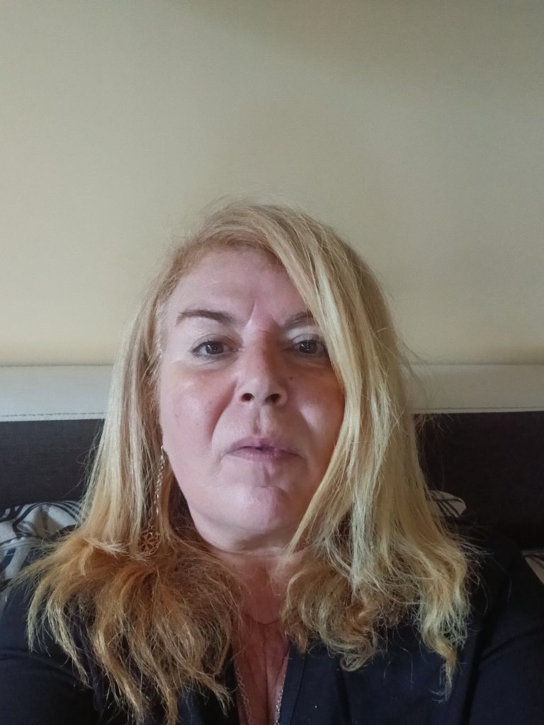 Blonde middle aged woman, light skinned, with dark-colored eyes and small earrings and a black top, seated on a couch. 