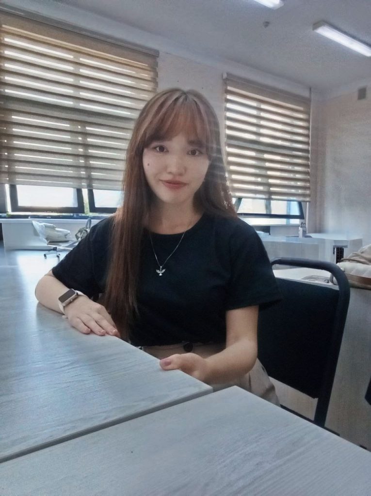 Young Central Asian woman seated in a classroom at a desk with long straight dark hair. She's wearing a black top and small star necklace.