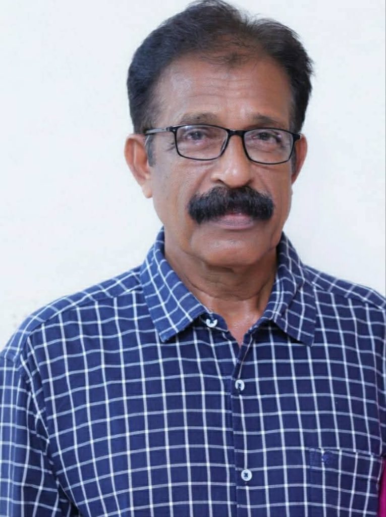 Older South Asian man in a blue and white checkered shirt, dark hair, and reading glasses.