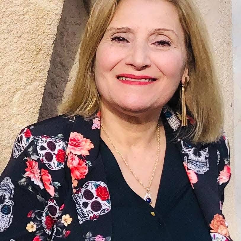 Middle aged light skinned woman with blonde hair, brown eyes, and a floral top and dangly earrings.