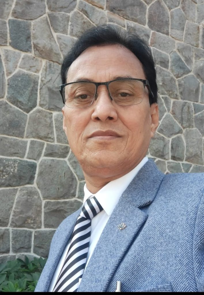 Older middle aged South Asian man with short dark hair and reading glasses  and a gray suit and tie standing in front of a stone wall. 