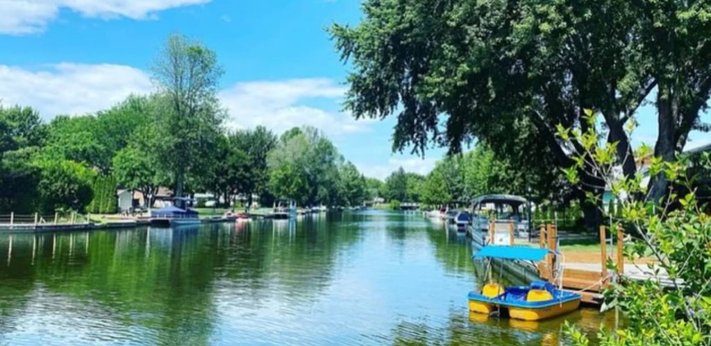 Calm water on a sunny day with some green trees and small boats by the shore. 