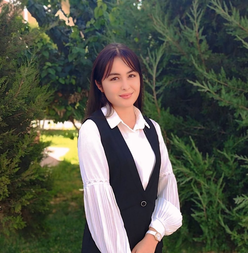 Young Central Asian woman in a white top and black vest with long dark hair standing in front of a few juniper bushes on a lawn. 