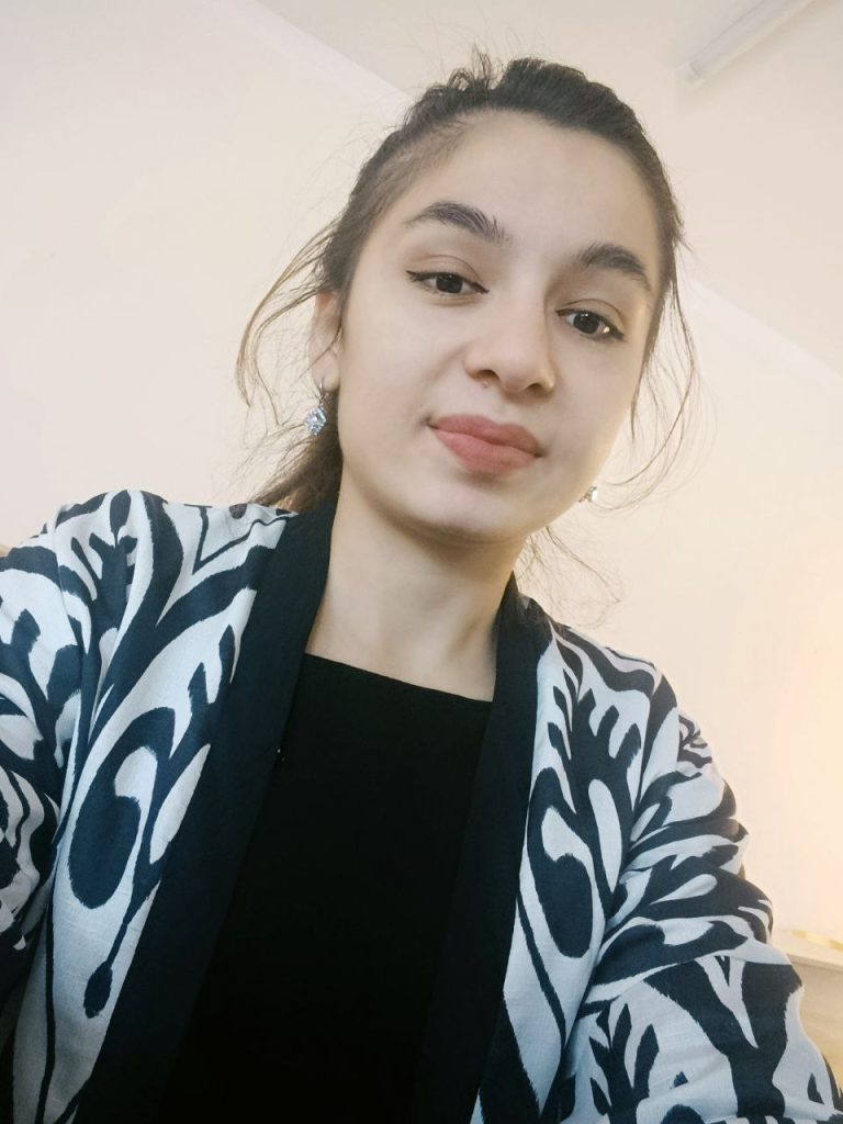 Young Central Asian woman with dark hair, brown eyes, and a black and white patterned sweater over a black top.