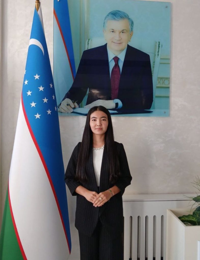 Young Central Asian woman in a black coat and dress pants and long dark hair standing beneath a photo of a male historical figure and the Uzbek flag.