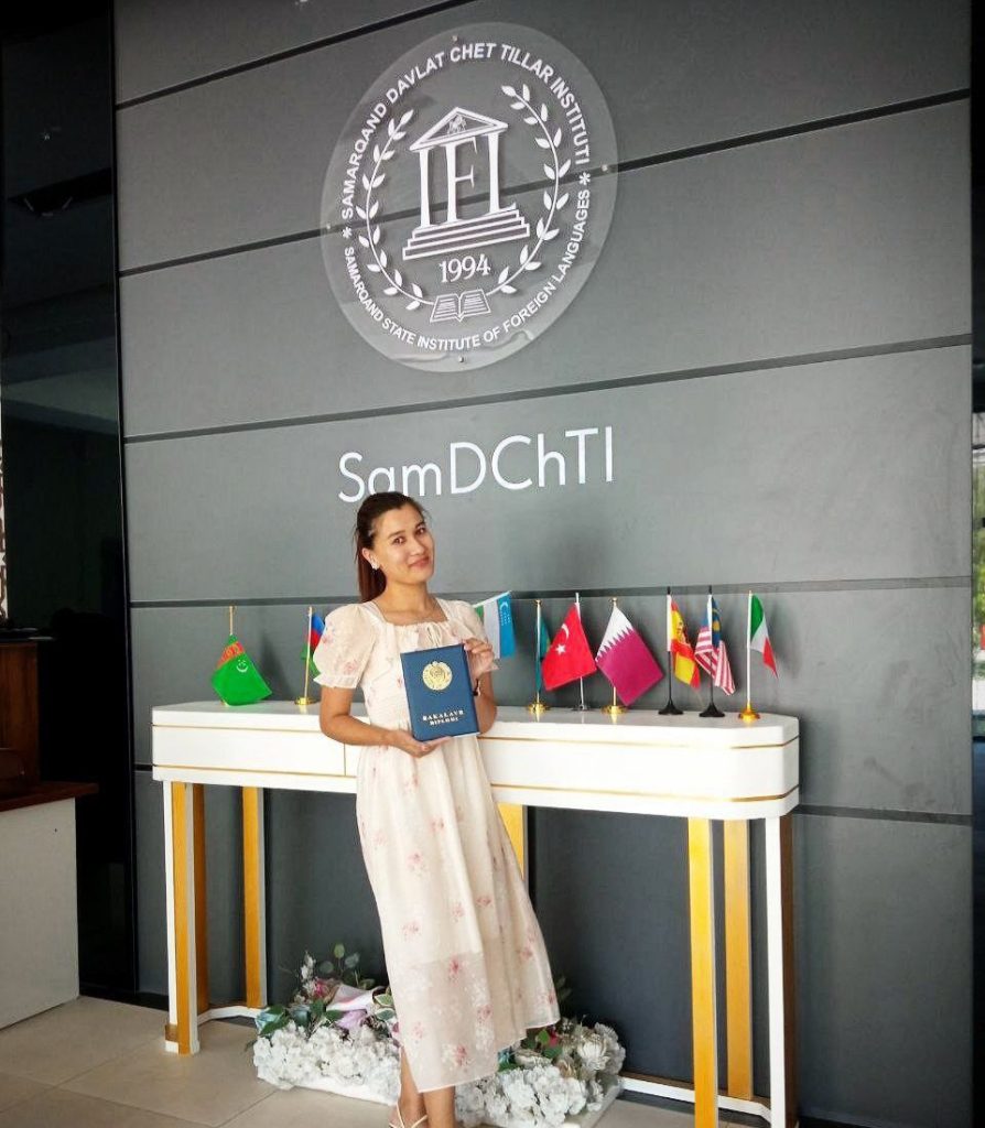 Young Central Asian woman in a classroom in front of a gray wall in a white dress with tiny pink flowers. She holds a certificate in front of a bunch of flags.