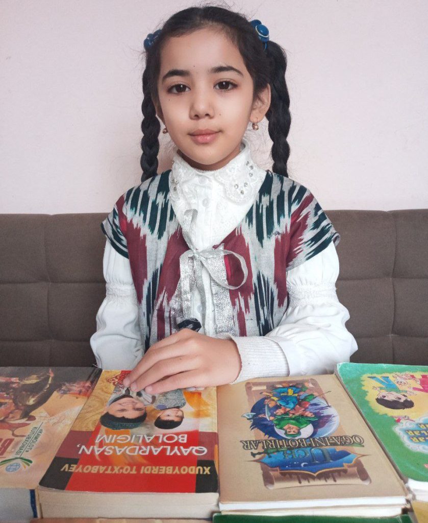 Young Central Asian girl with a patterned vest over a white top and two dark braided pigtails, seated on a couch in front of a table with some books.