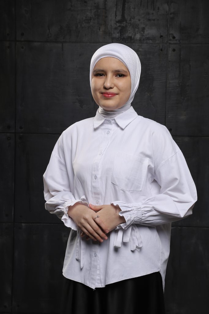 Central Asian woman in a white headscarf and collared shirt and black skirt. Her hands are folded in front of her chest. 