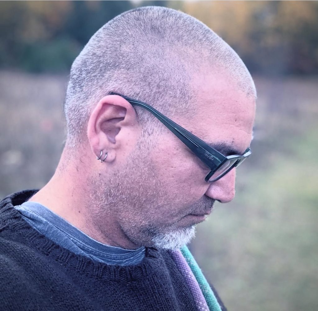 Middle aged almost bald white man with reading glasses and a sweater over a blue shirt and two earrings. 