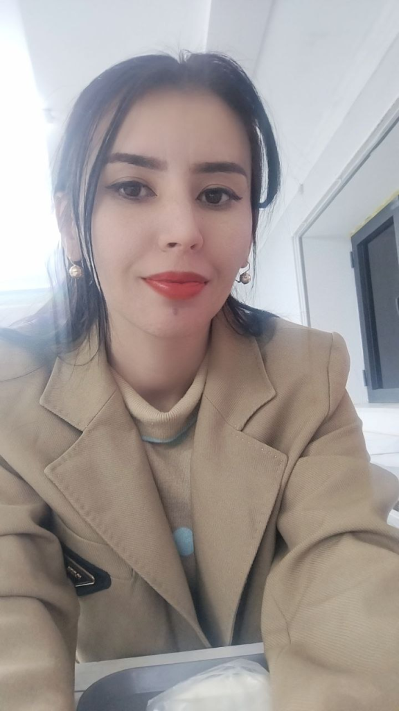 Young Central Asian woman with straight dark hair and a brown coat and small white pearl earrings. 