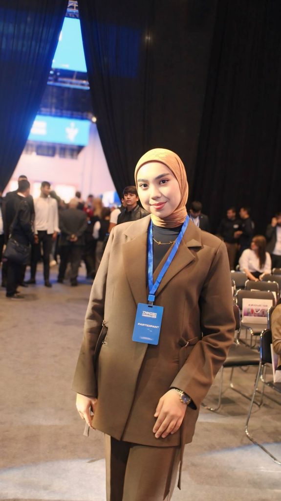 Young Central Asian woman with a tan coat and headscarf and blue lanyard at a convention near a blue curtain.