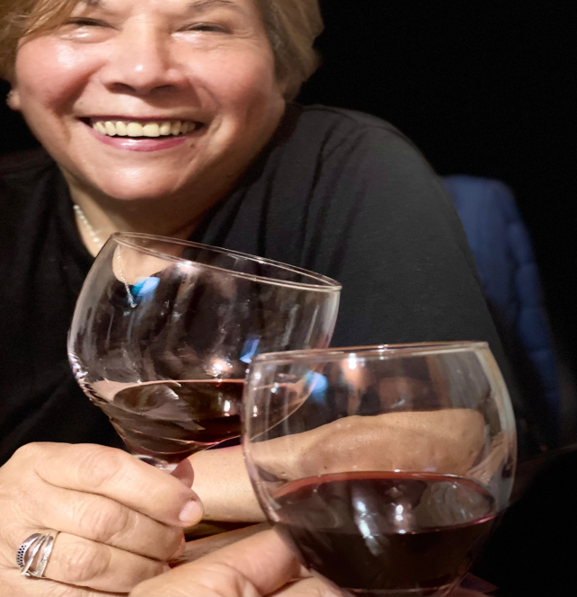 Middle aged smiling white woman with short blonde hair, holding two glasses of red wine.