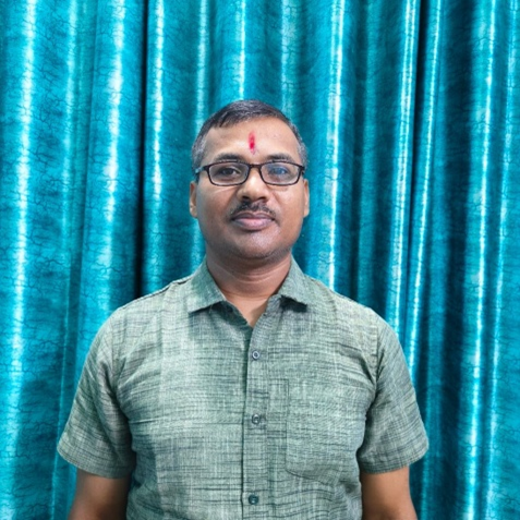 Middle aged South Asian man in a collared striped green shirt in front of a blue curtain.