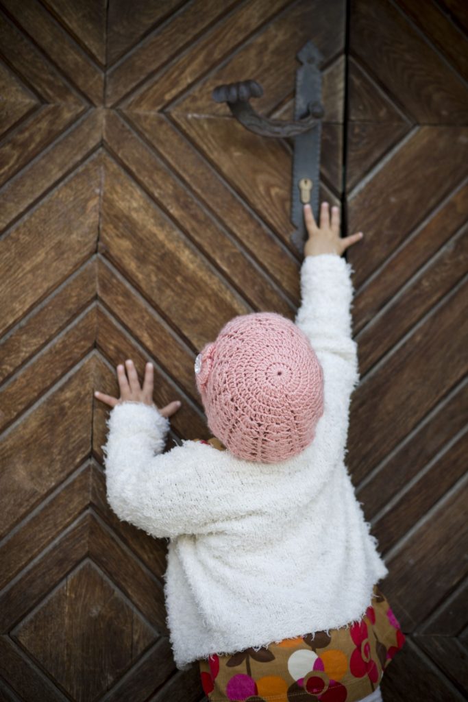 Small child in a pink knit hat and white coat and flowered dress trying to open a wooden paneled door. 