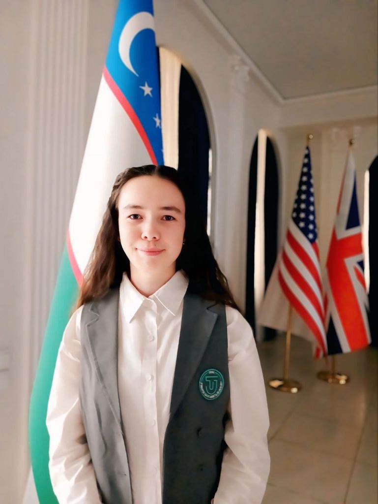 Young Central Asian woman with dark curly hair, a gray vest, and white collared shirt, standing in a room of large flags: British, Uzbek, US. 