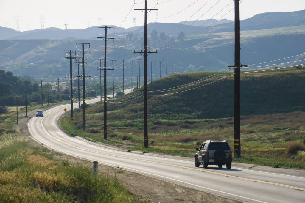 A few cars making their way down a curve in a country road near green grassy hills and trees and telephone poles. 