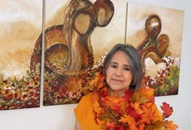 Older middle aged Latina woman with garland of fall leaves and a yellow top, standing in front of brown and red and green artwork.