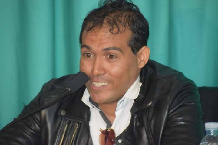 Young middle aged Arab man with dark curly hair and brown eyes. He's in a black jacket and white shirt.