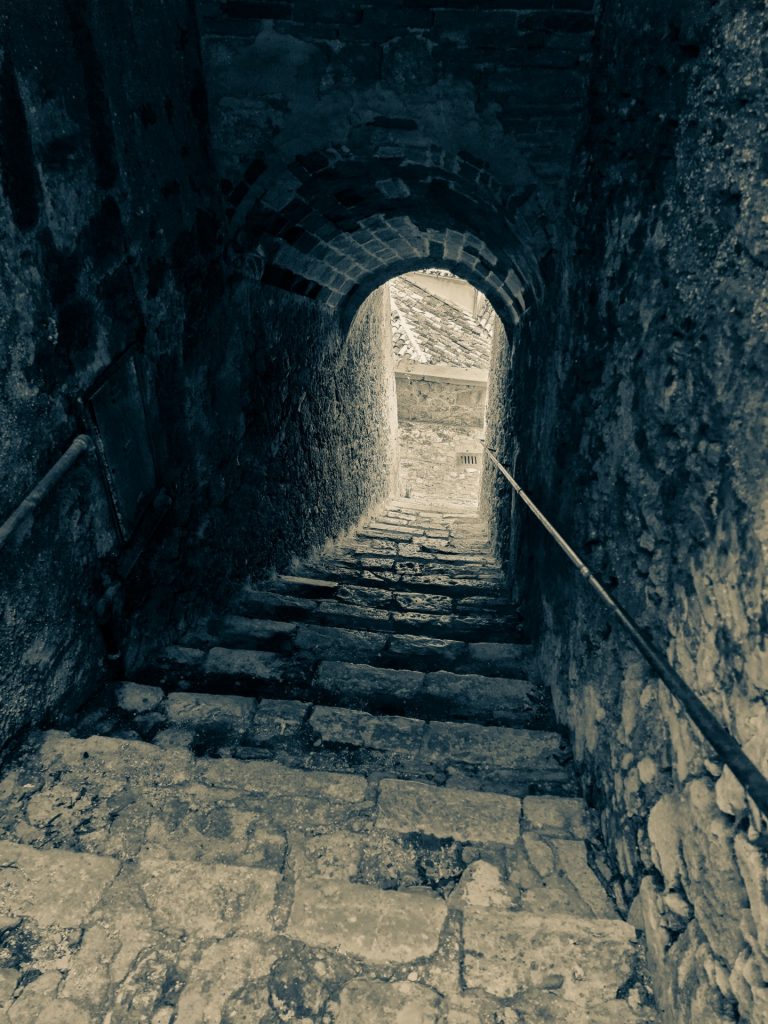 Old weathered stone steps in a descending path in an old castle. Light at the end of the tunnel, curved door. 