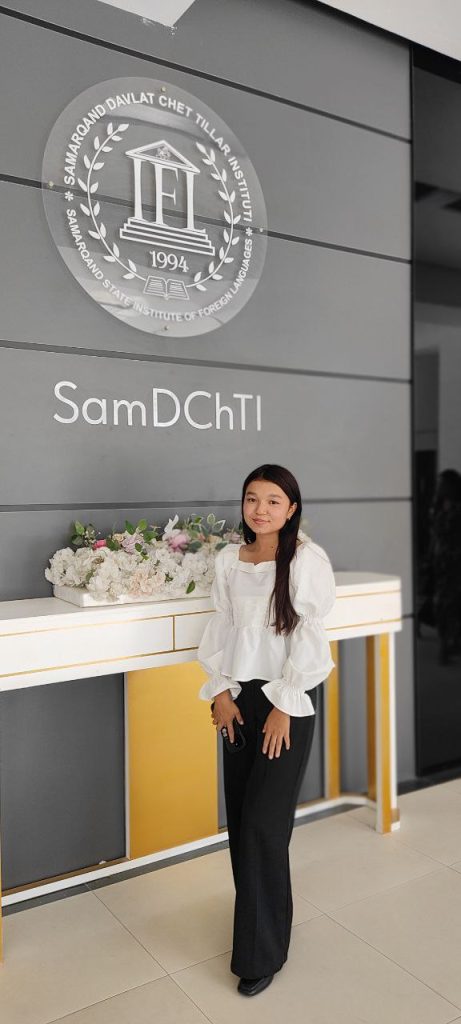 Young Central Asian woman with long dark hair, a white ruffled blouse, and dark pants. She's standing near flowers indoors at an event. 