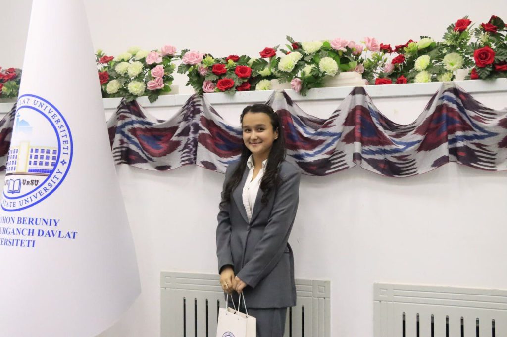 Young Central Asian woman with long curly dark hair, a white blouse, and gray coat and dress pants, on stage near a banner and flag and with flowers.