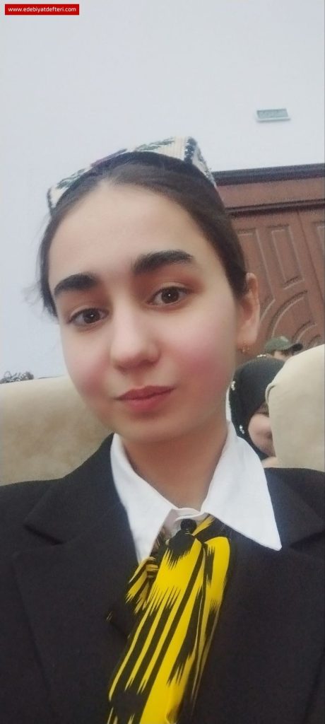 Young Central Asian woman with an embroidered headdress, dark straight hair up in a bun, brown eyes, a black coat, white collared shirt, and a black and yellow tie. 