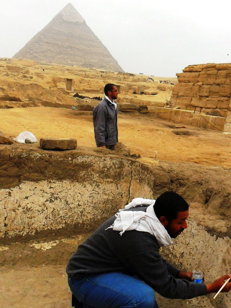 Two middle aged men survey an archaeological site in the desert on a sunny but hazy day. Both are in jackets and white scarves. Rocks nearby are stacked into structures.