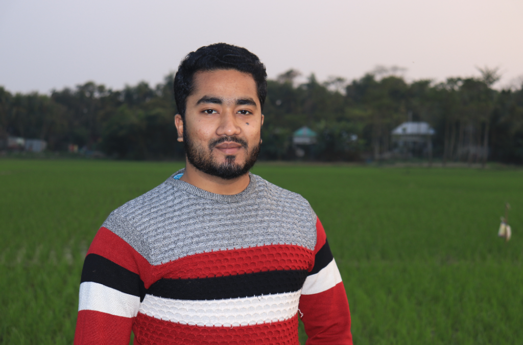 Middle aged South Asian man with short dark hair, beard, and mustache and a gray, black, white and red sweater standing in a field of grass at dawn or dusk. 