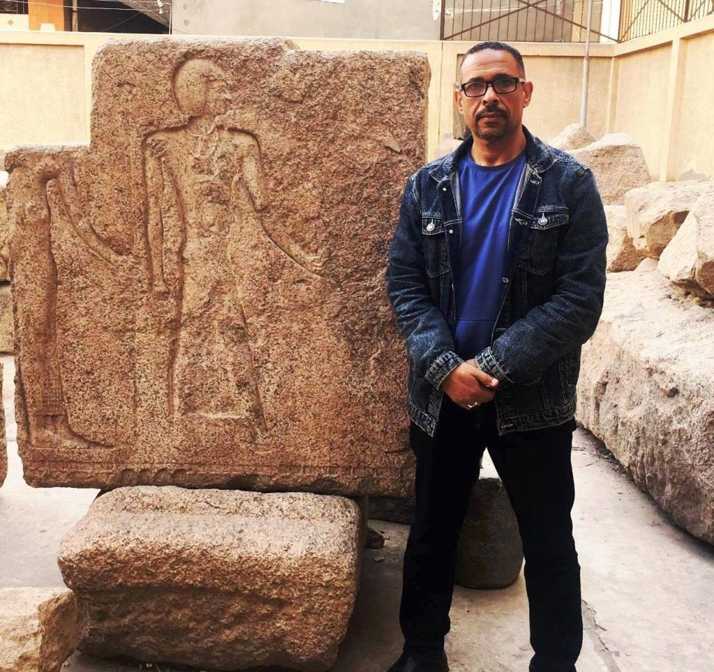 Middle aged Egyptian man with a jean jacket over a blue tee shirt. He's got reading glasses and stands in front of a rock with an ancient carving.