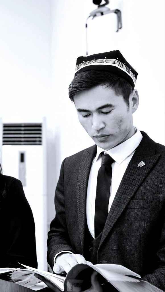 Black and white photo of a Central Asian teen boy in a black suit coat, white collared shirt, and dark tie. He's looking down and has an embroidered headdress on his head.