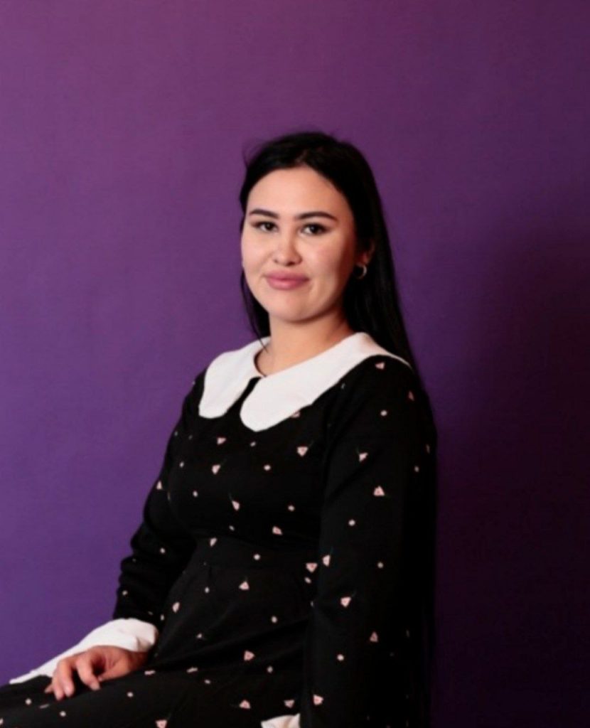 Young Central Asian woman with long dark straight hair and a black dress with white spots and a white collar, purple background.