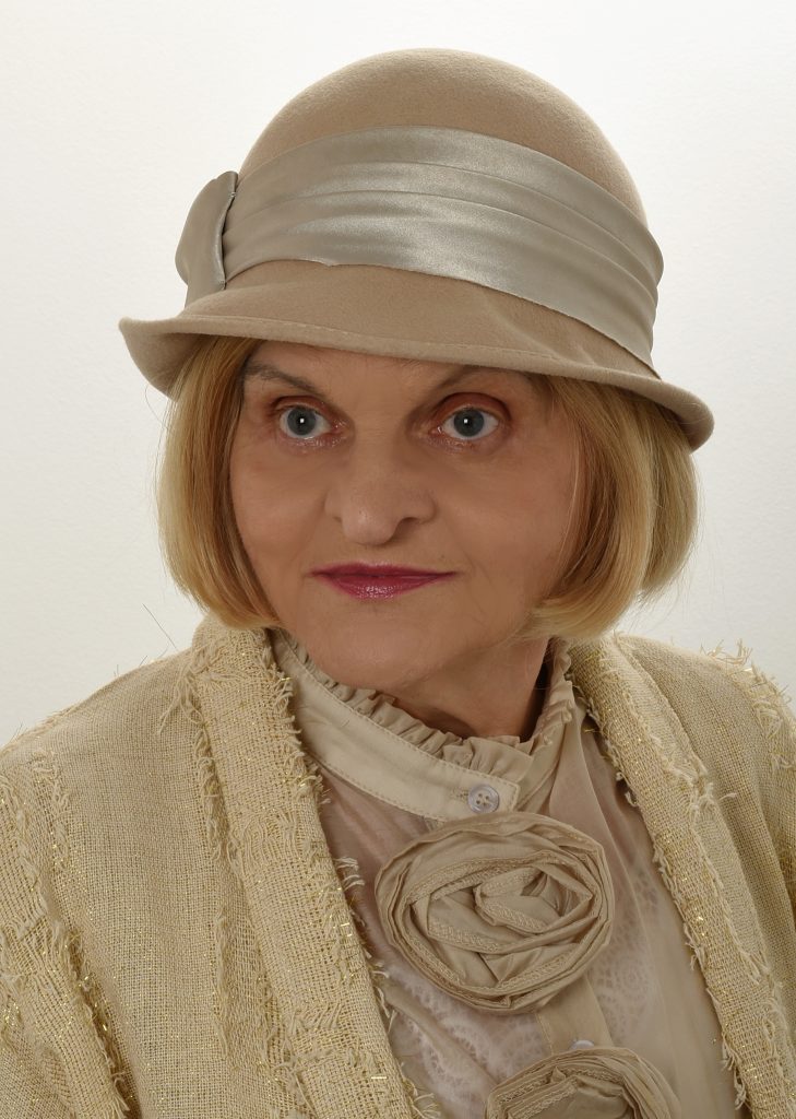 Young European woman with short blonde hair, blue eyes, and a light tan ruffled top and coat and a tan hat with a ribbon. 