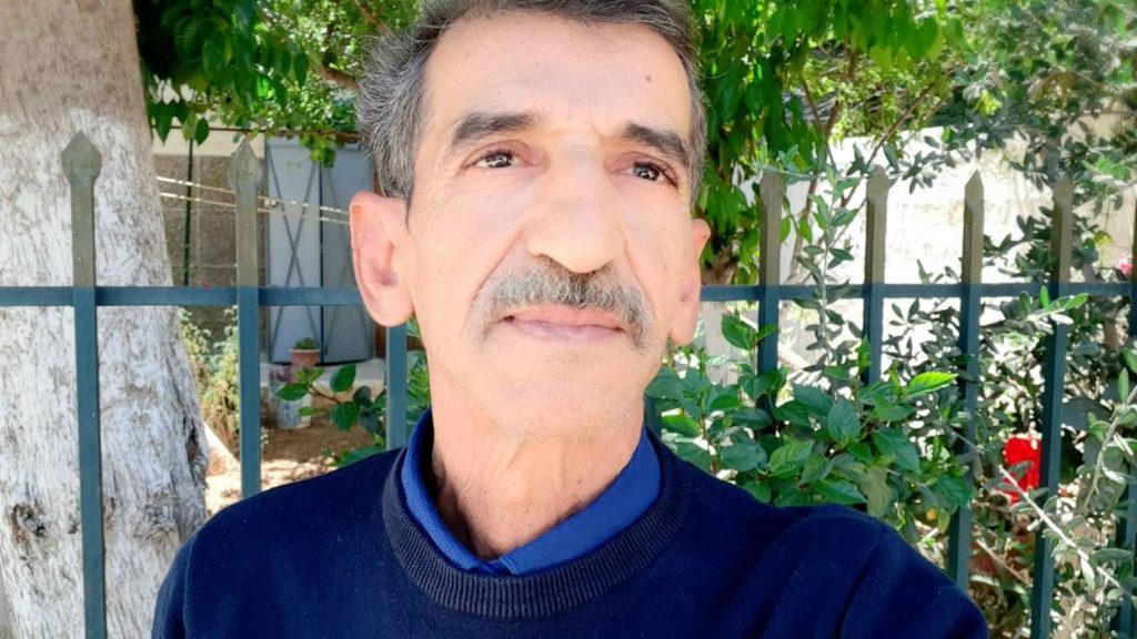 Older middle aged man with gray hair and a mustache and a dark blue sweater outside in a garden with blooming plants behind him.