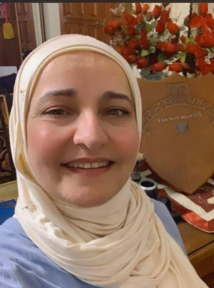 Smiling light skinned Arab woman with a white headscarf and a blue top standing near a bouquet of roses.