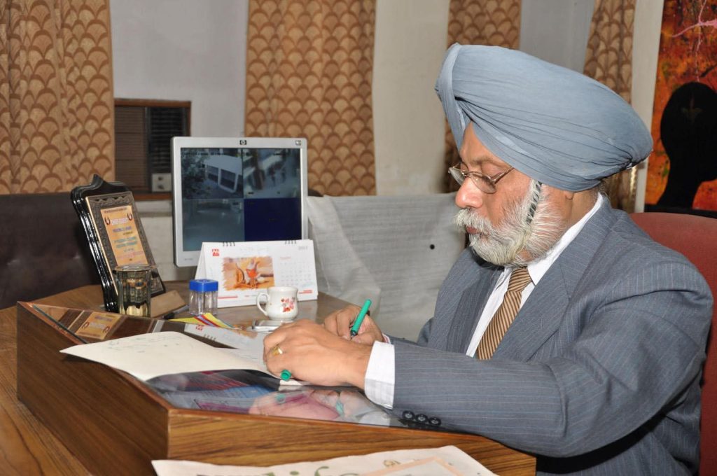Older South Asian man with a white beard and mustache and gray turban and coat and brown tie and white shirt seated at a desk with papers and a computer. 
