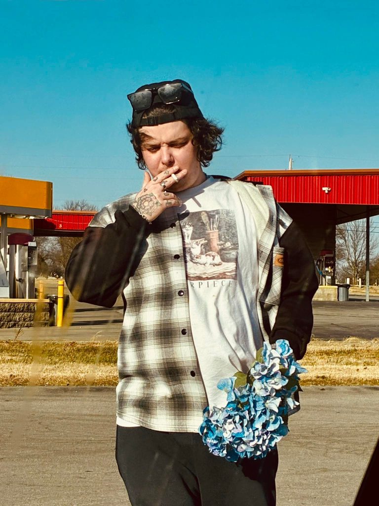 Young white man smoking a cigarette. He's got curly black hair and a baseball cap and a plaid shirt with buttons over a shirt with some design on it. He's at a gas station on a sunny day and is holding blue hydrangeas.