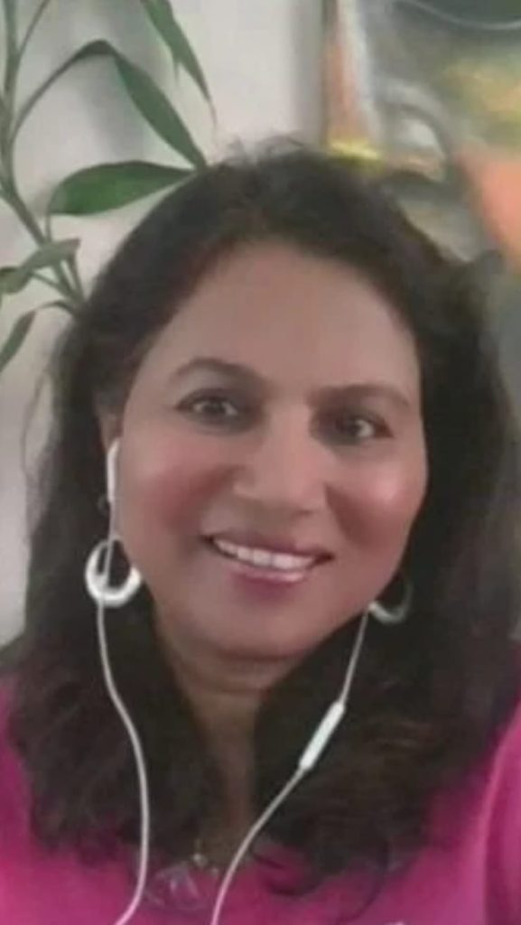 Young middle-aged South Asian woman with short dark curly hair, earrings, earphones, and a pink top, seated next to a plant .
