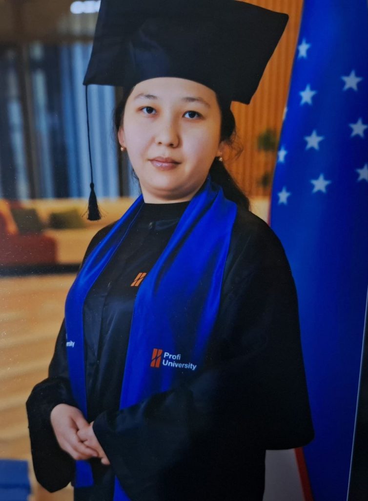 Young Central Asian woman with long dark hair and earrings in a blue and black graduation cap and gown standing on stage next to a flag. 
