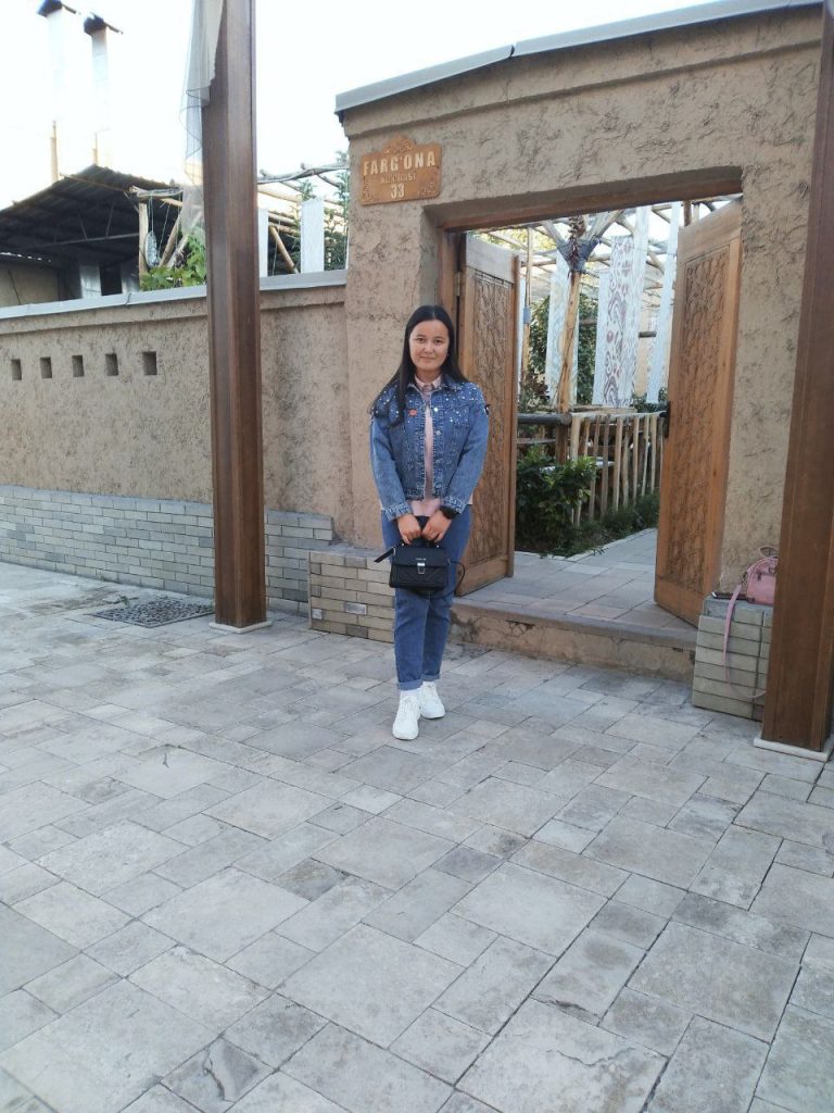 Young Central Asian girl in a jean jacket, light colored blouse, and jeans and white tennis shoes. She's holding a black purse and has long dark hair. She's in front of an entrance to a stone building with trees in a courtyard. 