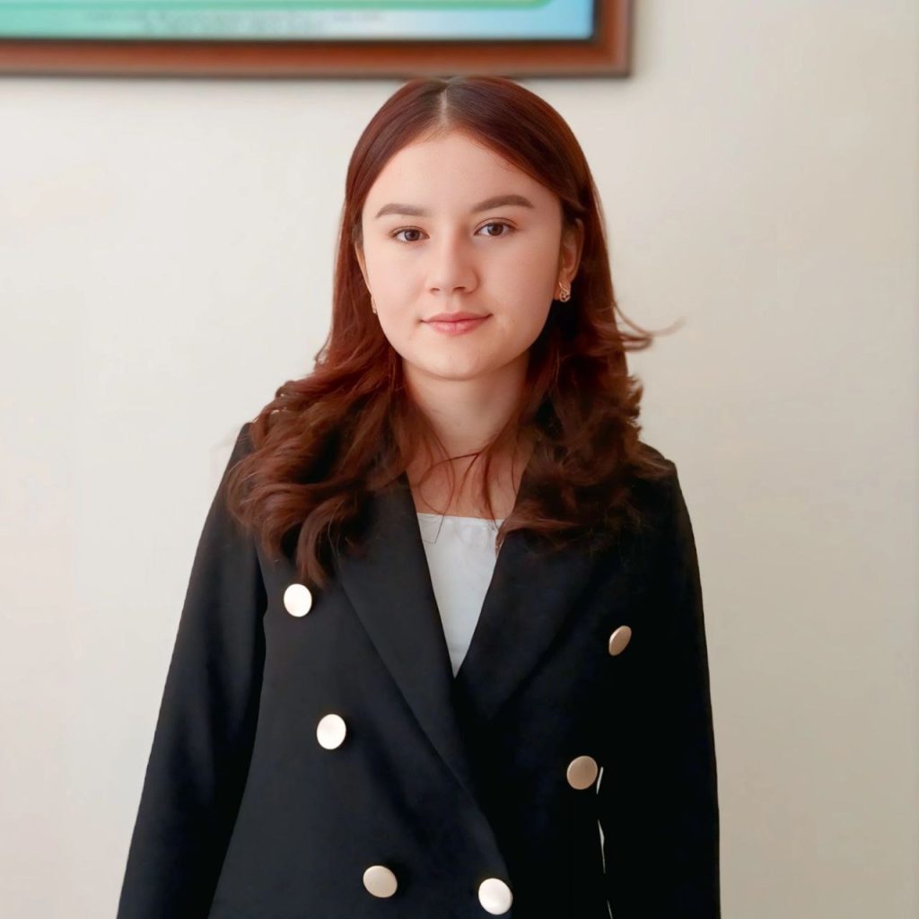 Young Central Asian woman with curly dark hair, brown eyes, and a black coat with large buttons and a white undershirt and small earrings. 