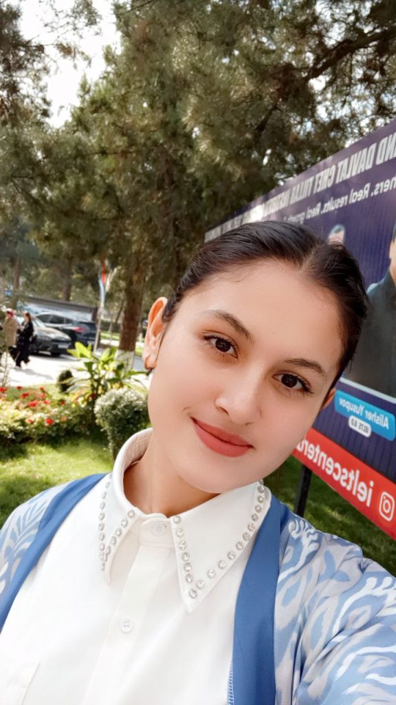 Young Central Asian woman with long dark hair, brown eyes, and a white collared top with a light blue and white coat, standing outside on a lawn near some flowers. 