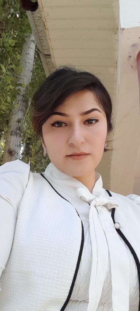Young Central Asian woman with straight dark hair in a bun, small earrings, and a white ruffled blouse with black trim, outside near a tree. 