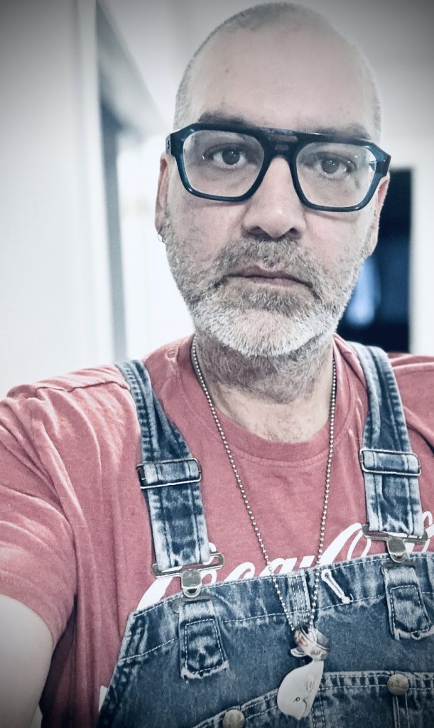 Middle aged white Canadian man, bald, trimmed mustache and beard, black glasses, with a red tee shirt and jean overalls.